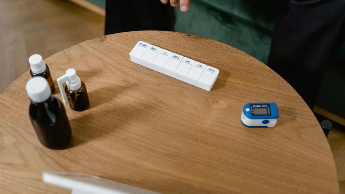 Weekly pill organizer, medicine bottles and a pulse oximeter on a wooden side table