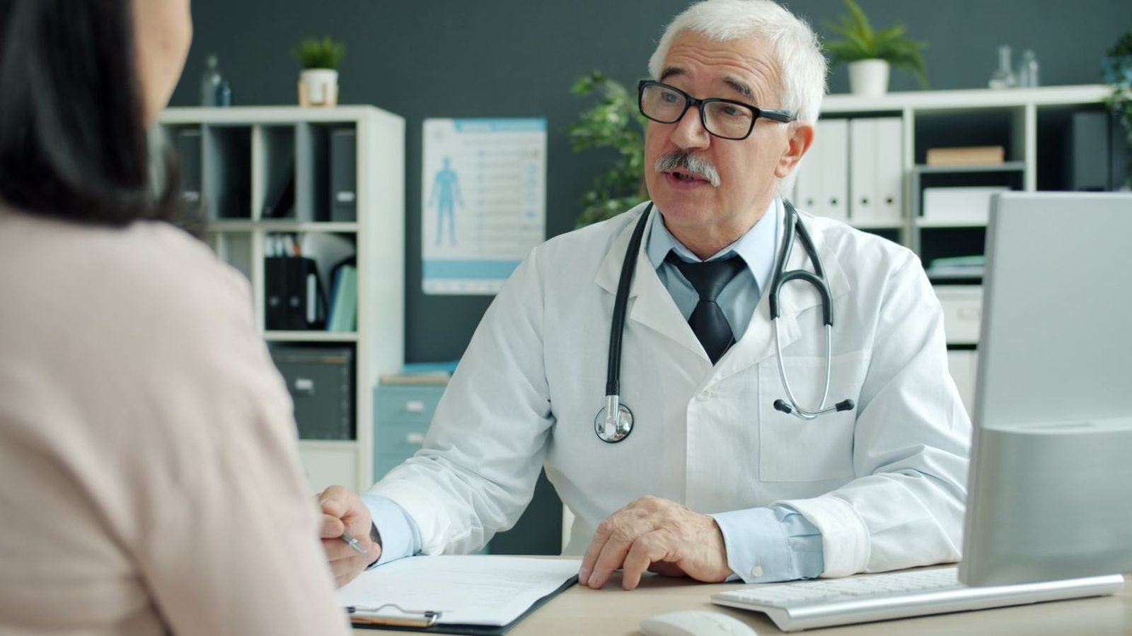A doctor in a white coat sitting across a desk in a consulting room, talking a patient through a decision with notes and a computer between them