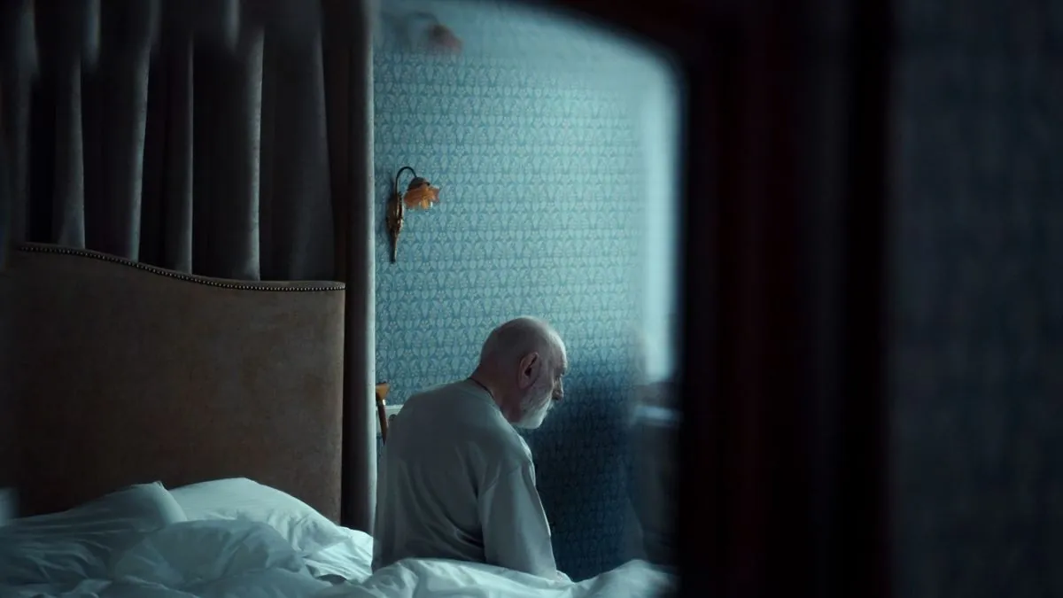 Older man sitting on the edge of his bed in a dimly lit bedroom at night, seen from behind